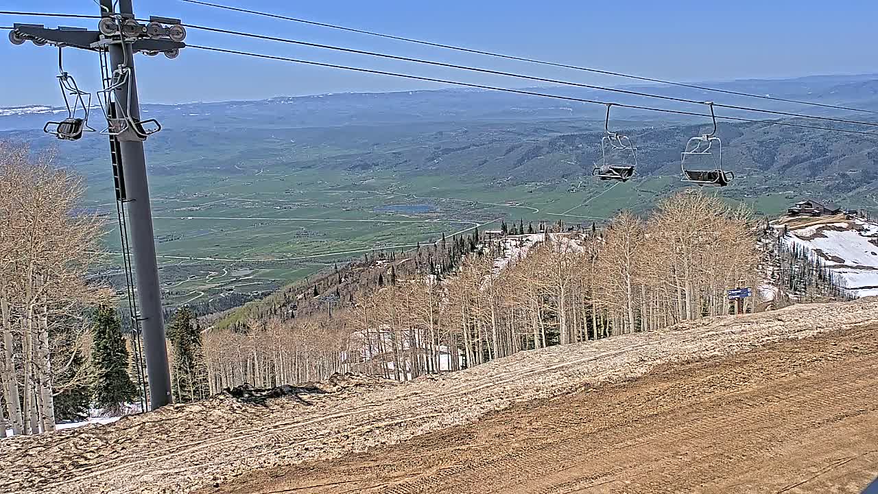 A ski lift with empty chairs overlooks a mountainous landscape with bare trees, patches of snow, and a valley under a clear blue sky.