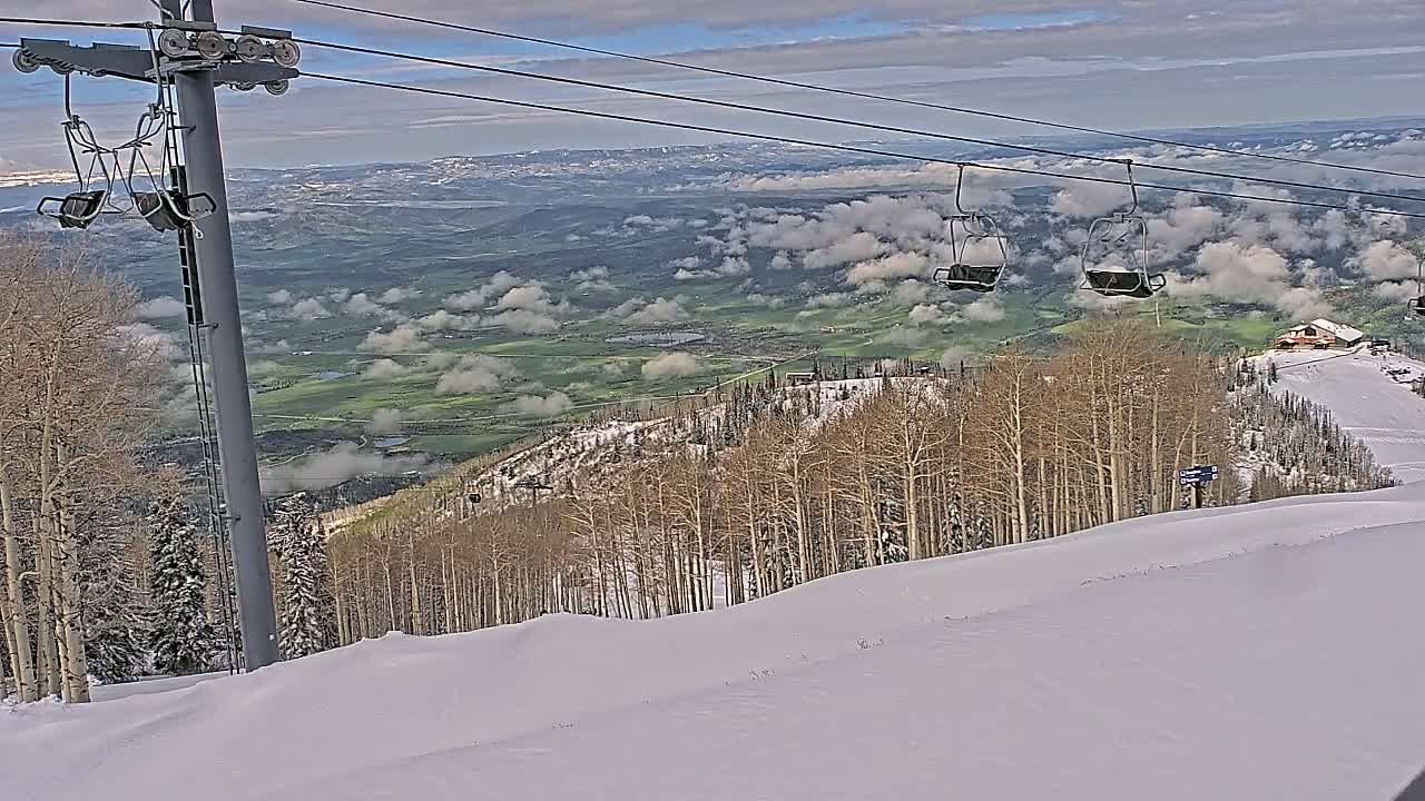 A snow-covered ski slope with a chairlift and a valley filled with clouds in the background under a partly cloudy sky.