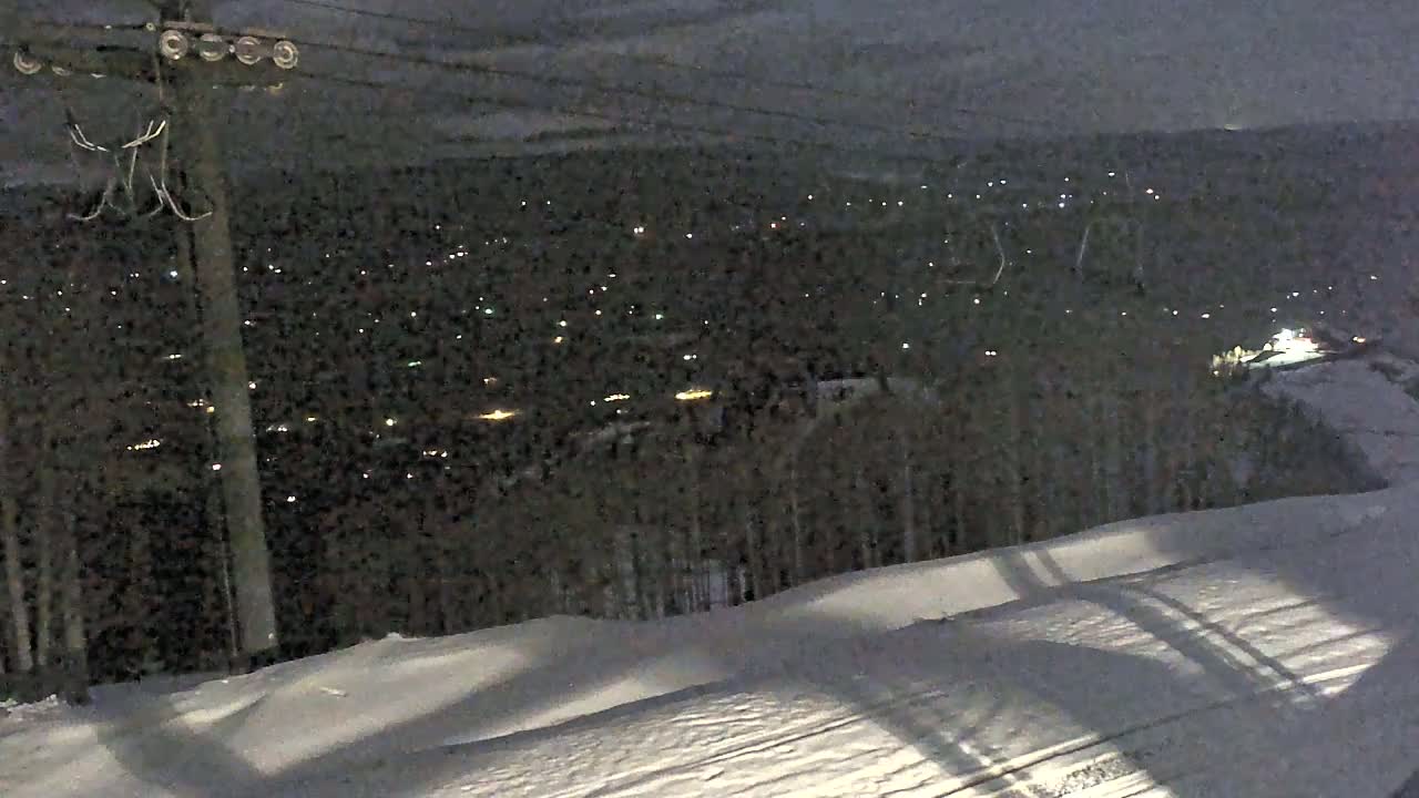 A snowy, nighttime landscape shows a distant town twinkling with lights below a hilltop vantage point.
