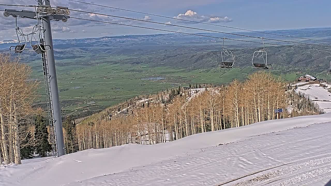 A snowy mountain slope with bare aspen trees, a chairlift, and a distant view of a green valley under partly cloudy skies.