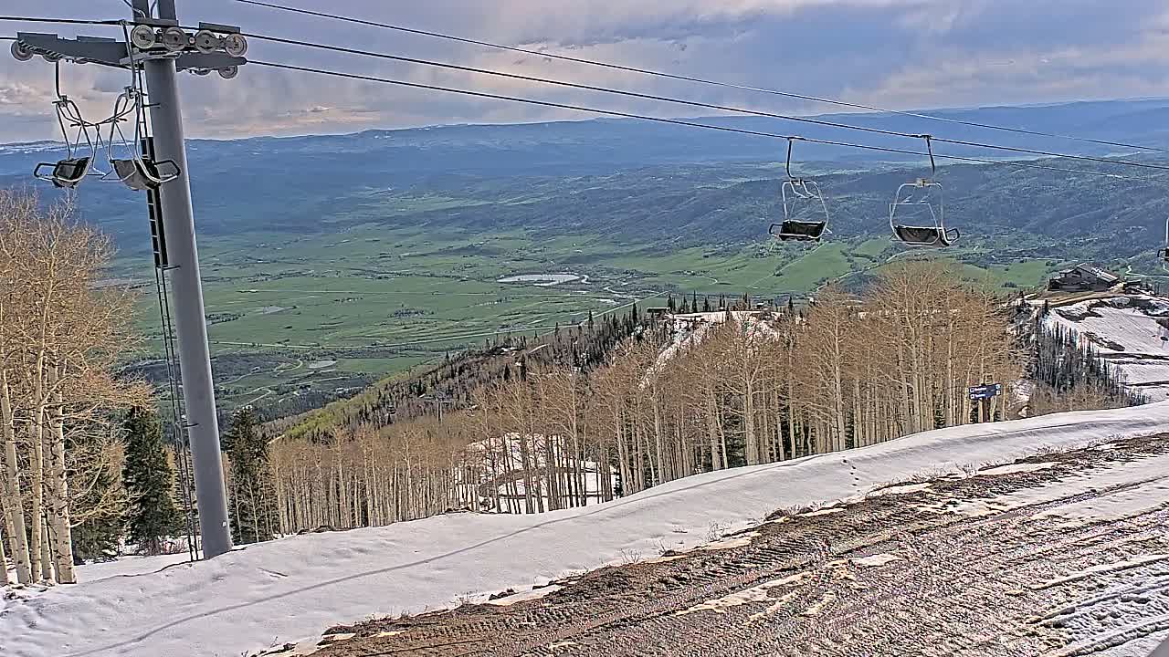 A chairlift ascends a snowy mountainside overlooking a vast green valley under a partly cloudy sky.