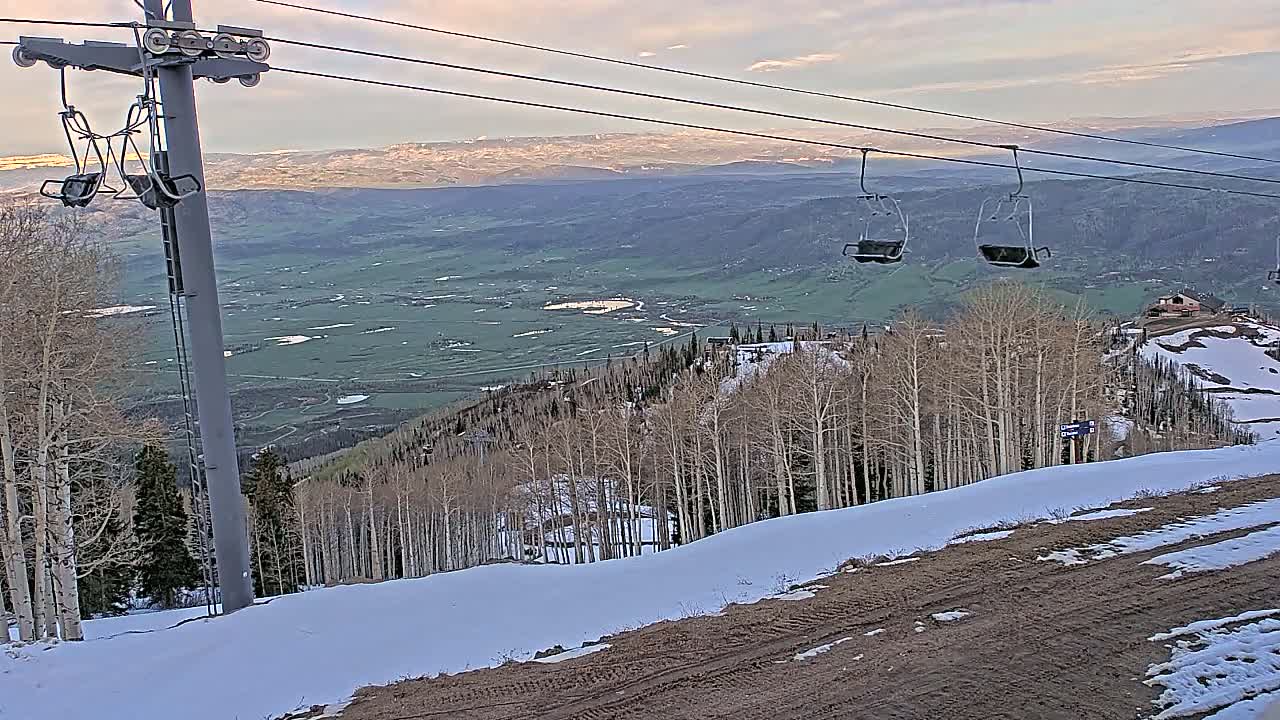 A ski lift with empty chairs overlooks a snow-dusted mountain slope and a vast valley under a partly cloudy sky.