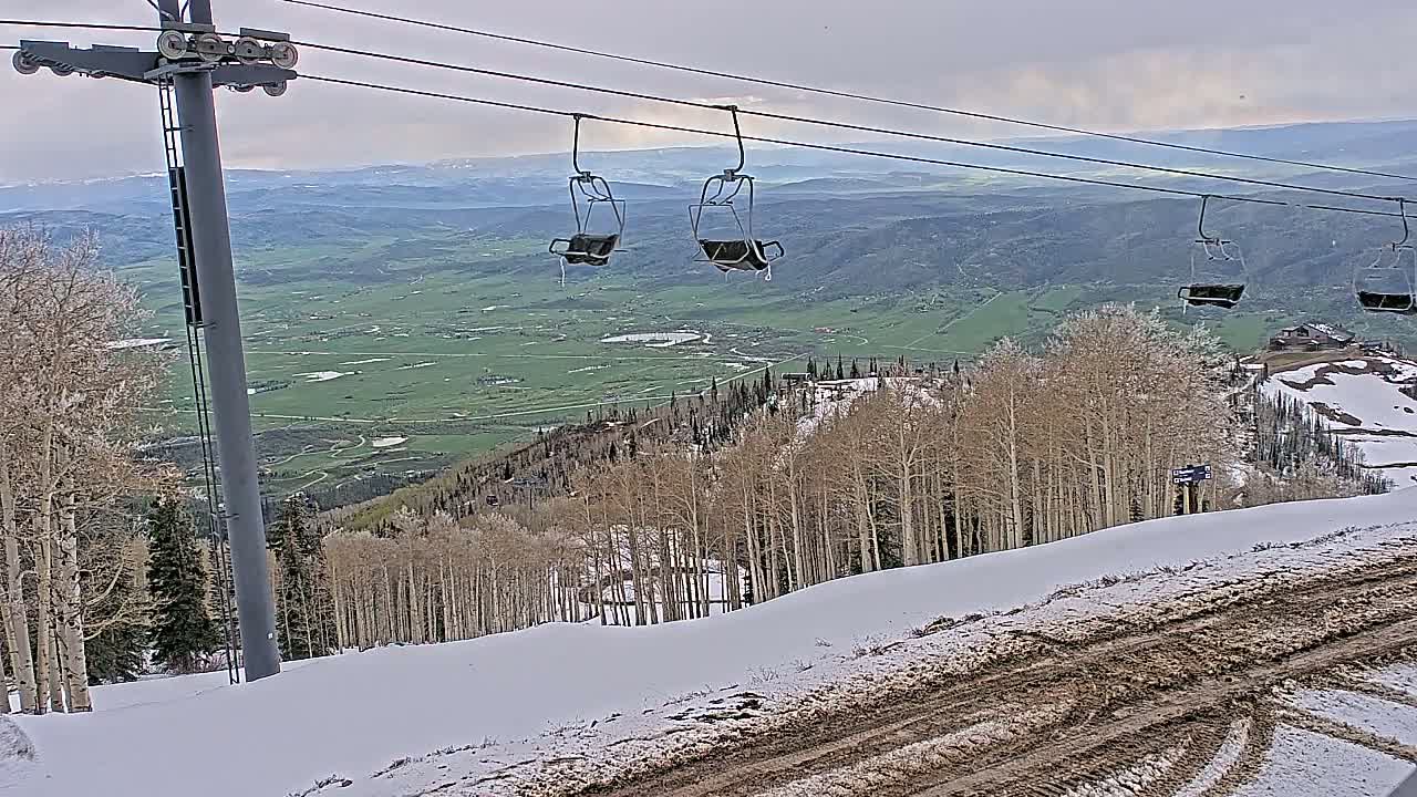 A chairlift ascends a snowy mountain slope overlooking a verdant valley under an overcast sky.