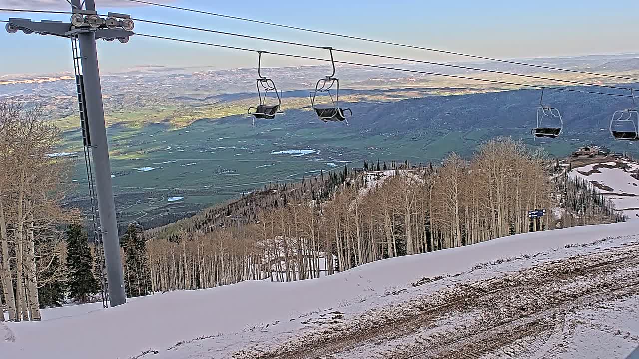A ski lift carrying empty chairs overlooks a snow-dusted mountain slope and a vast valley under a clear sky.