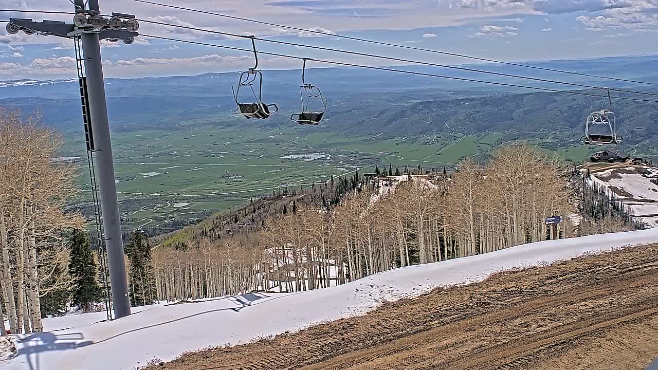 A ski lift with empty chairs overlooks a mountain valley under partly cloudy skies, with patches of snow remaining on the slopes and surrounding trees.