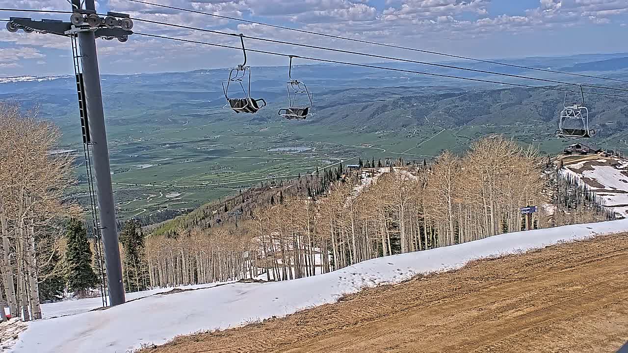 A chairlift ascends a mountain slope with patchy snow under a partly cloudy sky, overlooking a vast green valley.