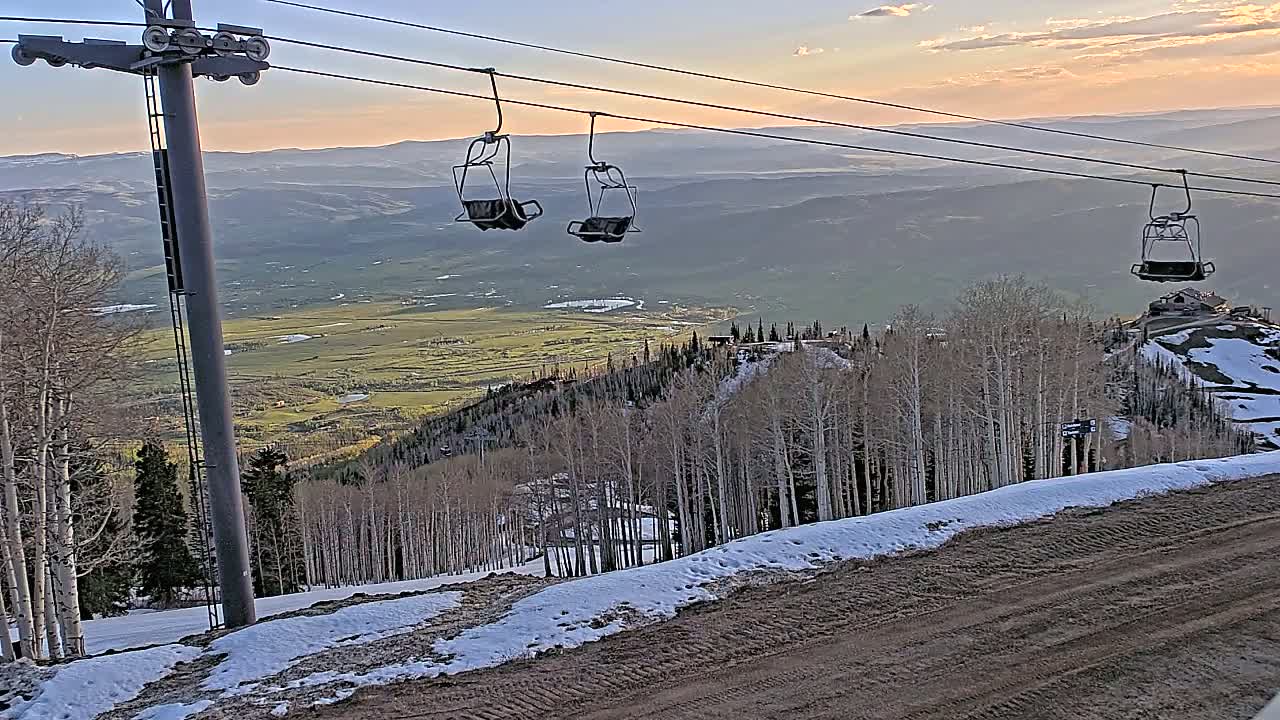 Three empty chairlifts hang from cables above a snow-dusted mountain slope overlooking a valley at sunset.