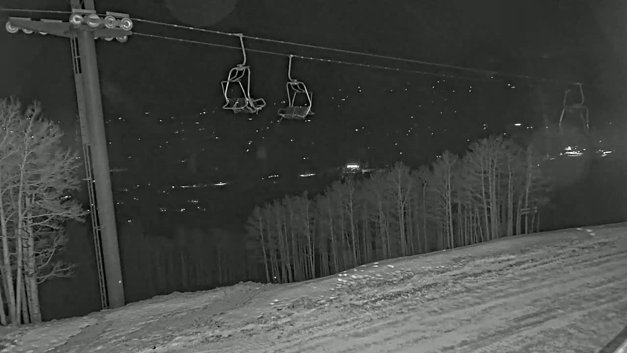 At night, a ski lift with empty chairs hangs over a snow-covered hill with leafless trees and distant lights.