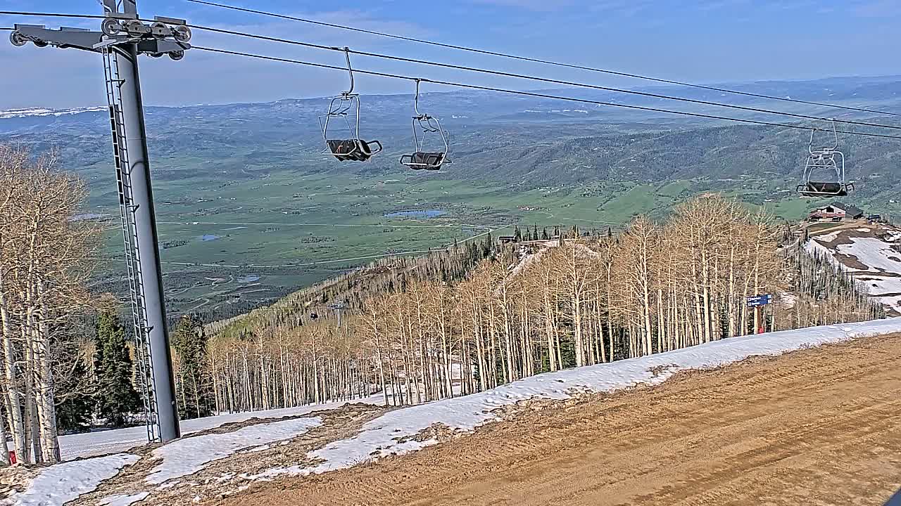 A mountaintop ski lift with empty chairs overlooks a valley of green fields and sparse, leafless trees under a clear blue sky.