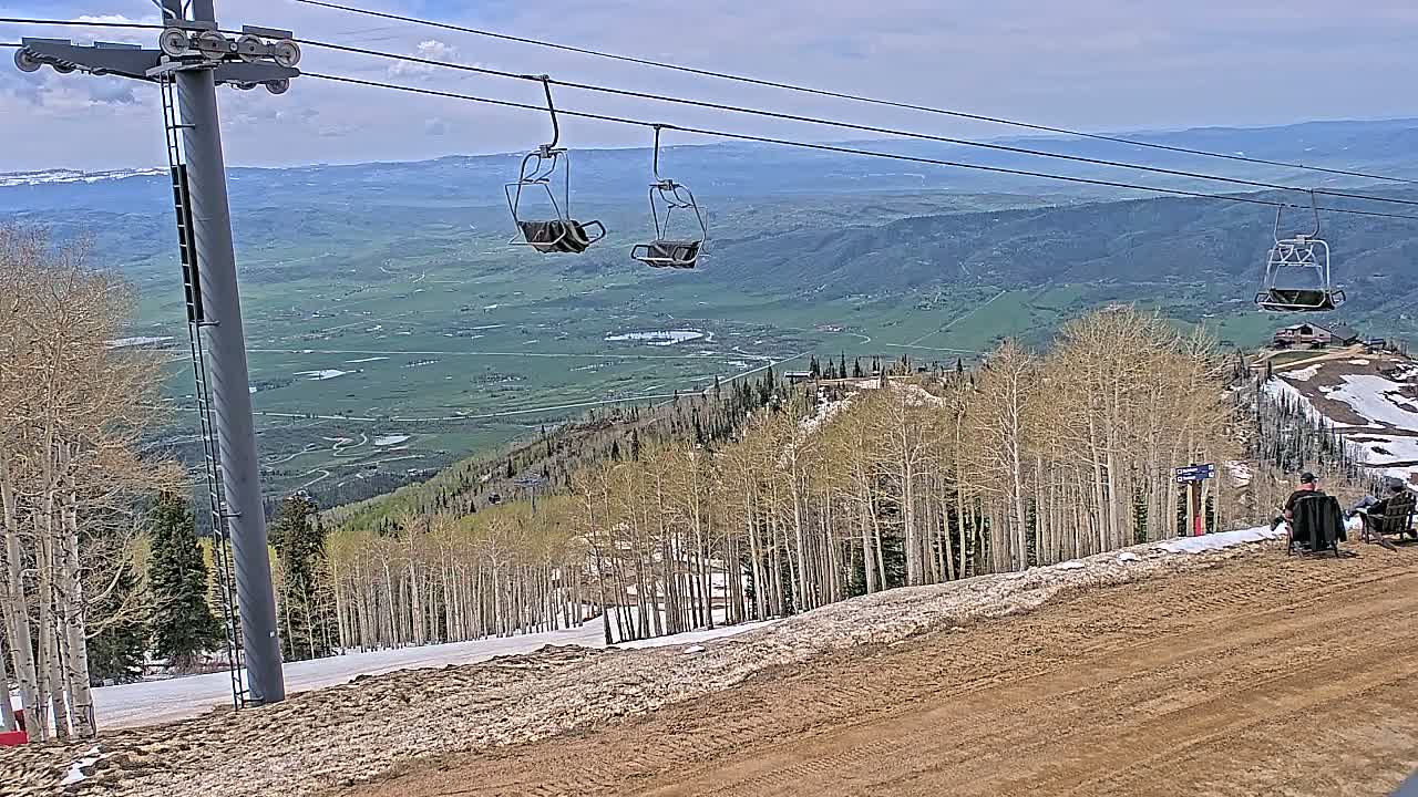 A mountaintop view shows a chairlift with empty chairs overlooking a vast green valley under a partly cloudy sky, with patches of snow and dirt on the mountainside and two people sitting on chairs in the foreground.