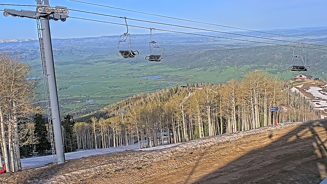 A ski lift with empty chairs overlooks a valley of green fields and a sparsely wooded mountainside with patches of snow under a clear blue sky.