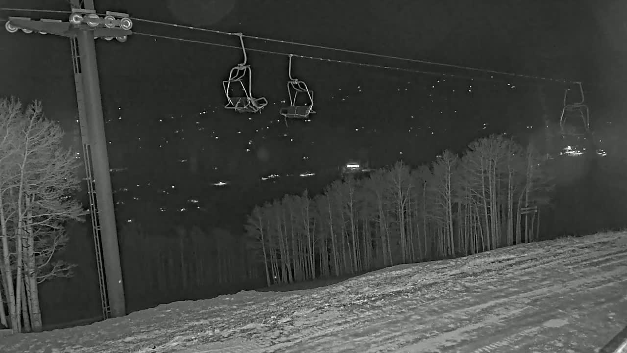 At night, empty chairlift chairs hang from a cable above a snow-covered slope and a dark forest dotted with distant lights.
