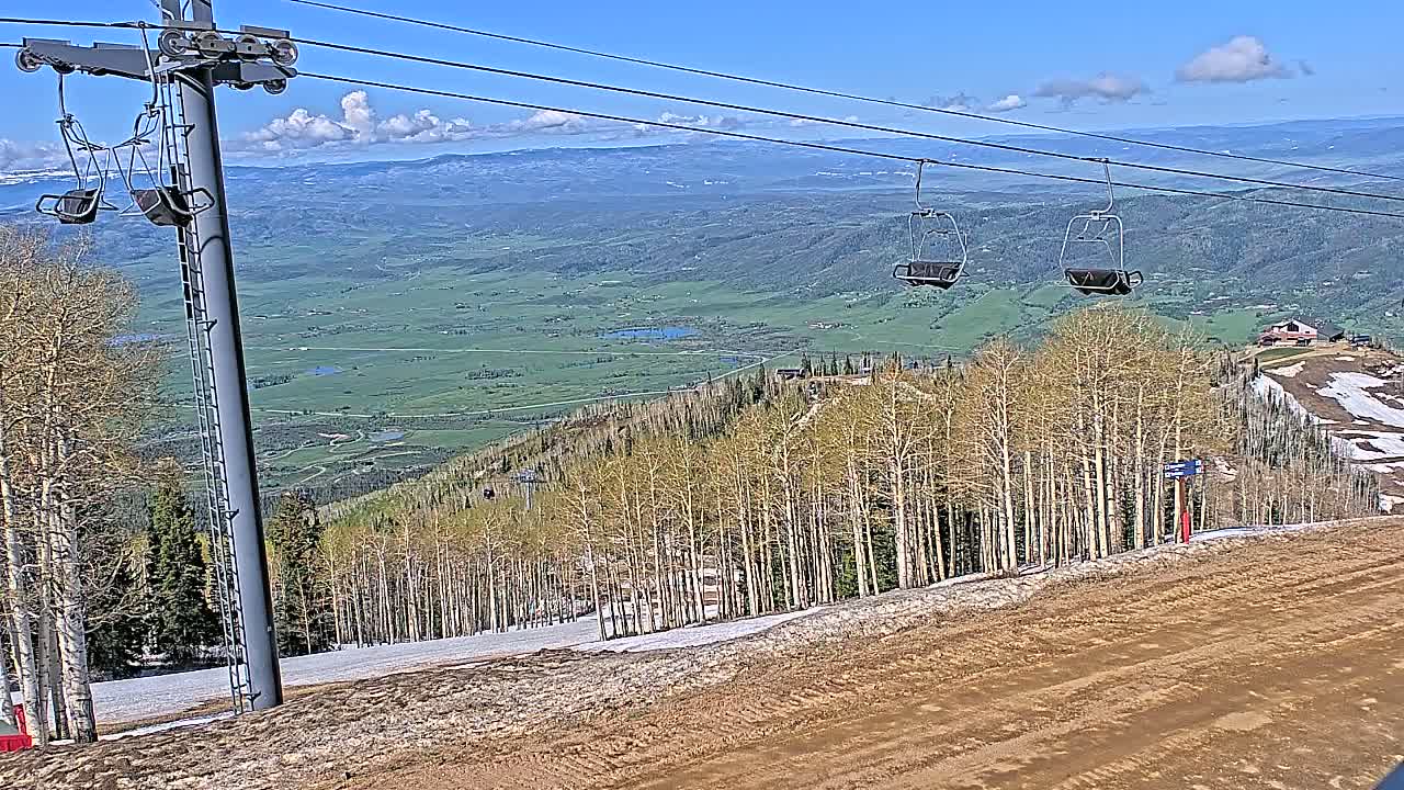 Two empty chairlifts hang above a mostly barren ski slope overlooking a vast green valley under a partly cloudy blue sky.