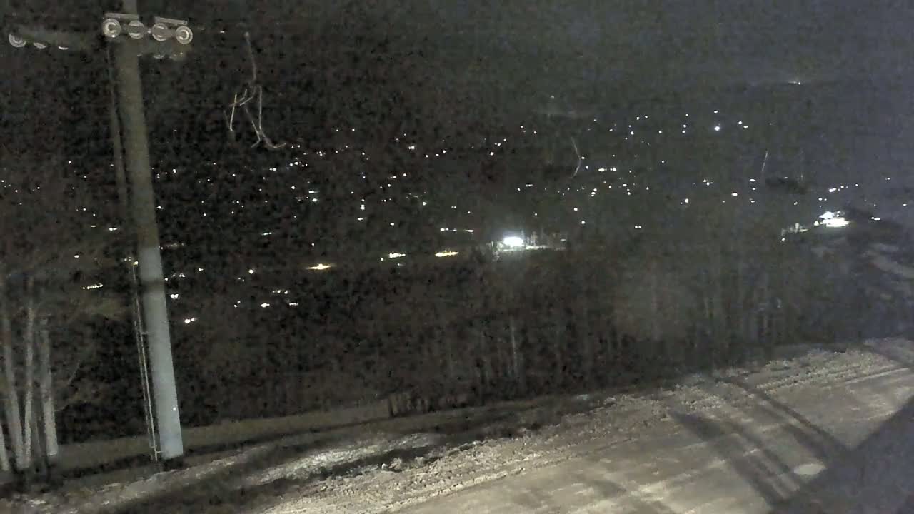 A nighttime view of a snowy, hilly landscape with distant lights from a town or village, and a ski lift pylon in the foreground.