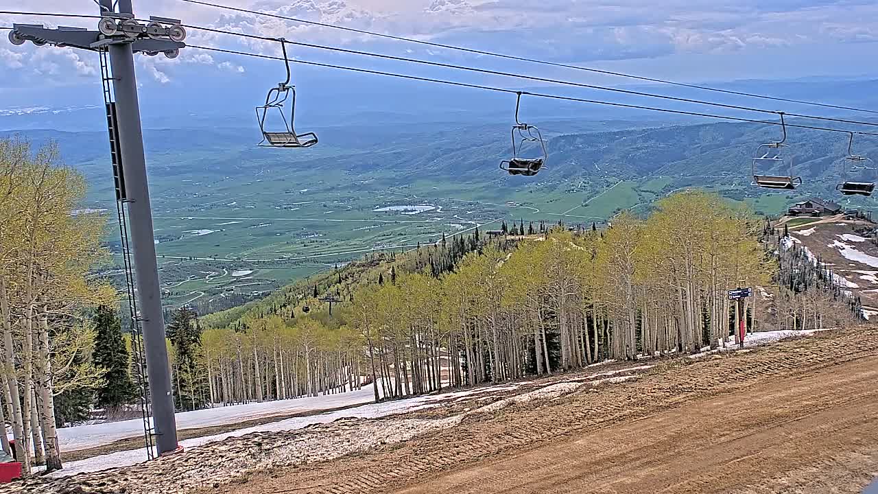 A chairlift ascends a mountain slope dotted with aspen trees, overlooking a vast green valley under a partly cloudy sky.