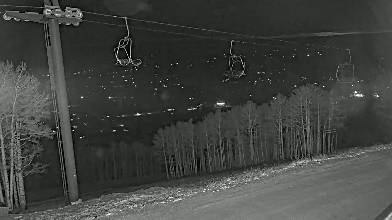 At night, empty ski lift chairs hang from cables above a snow-covered road and a dark forest overlooking a town illuminated by many distant lights.