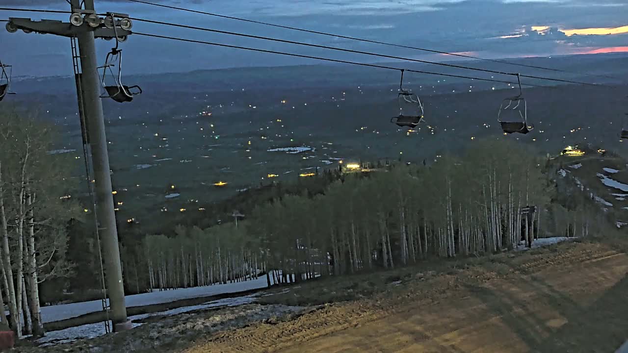 Empty chairlifts ascend a mountain at twilight, overlooking a valley speckled with distant lights and a snow-dusted ski slope below.