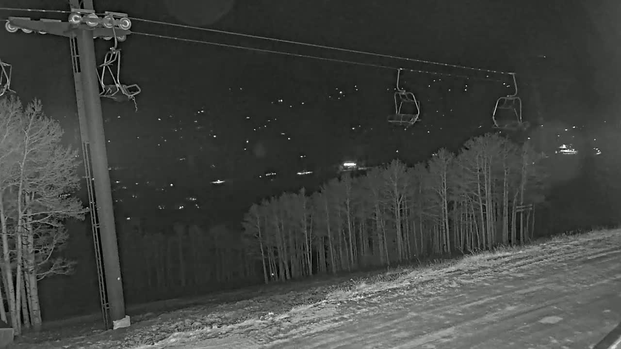 A nighttime view of a ski lift with empty chairs overlooking a snow-covered landscape and a distant town illuminated with lights.