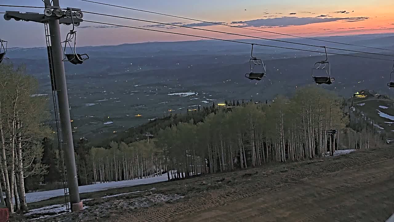 Empty ski lift chairs ascend a mountain at dusk overlooking a valley dotted with lights under a pinkish-orange sky.