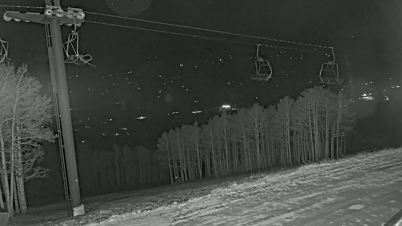 At night, a ski lift with empty chairs stands above a snow-covered slope and a line of bare trees, overlooking a distant town illuminated by lights.