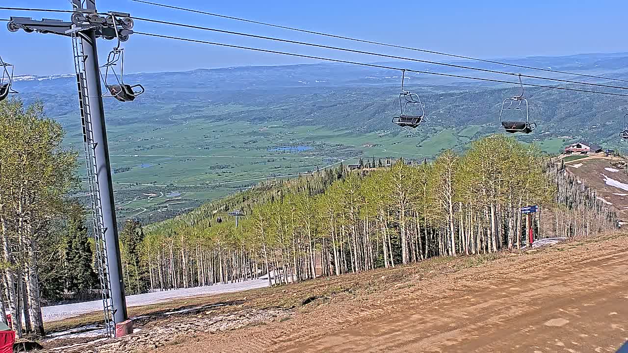 A ski lift with empty chairs overlooks a valley of green fields and aspen trees under a clear blue sky.
