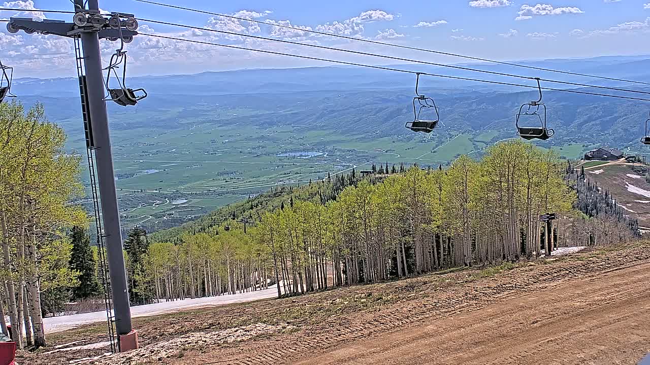 A mountain ski lift with empty chairs overlooks a vast green valley under a partly cloudy blue sky.