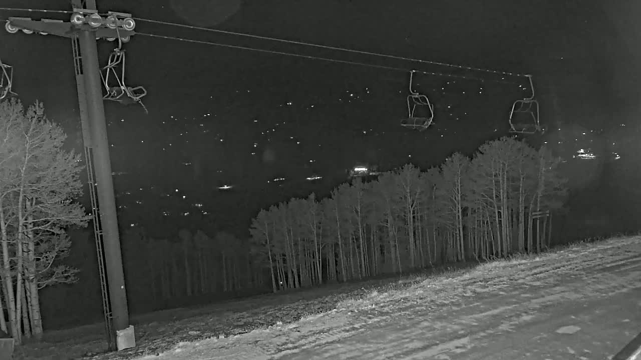 At night, a ski lift with empty chairs hangs over a snow-covered slope, beyond which are distant lights and a stand of trees.