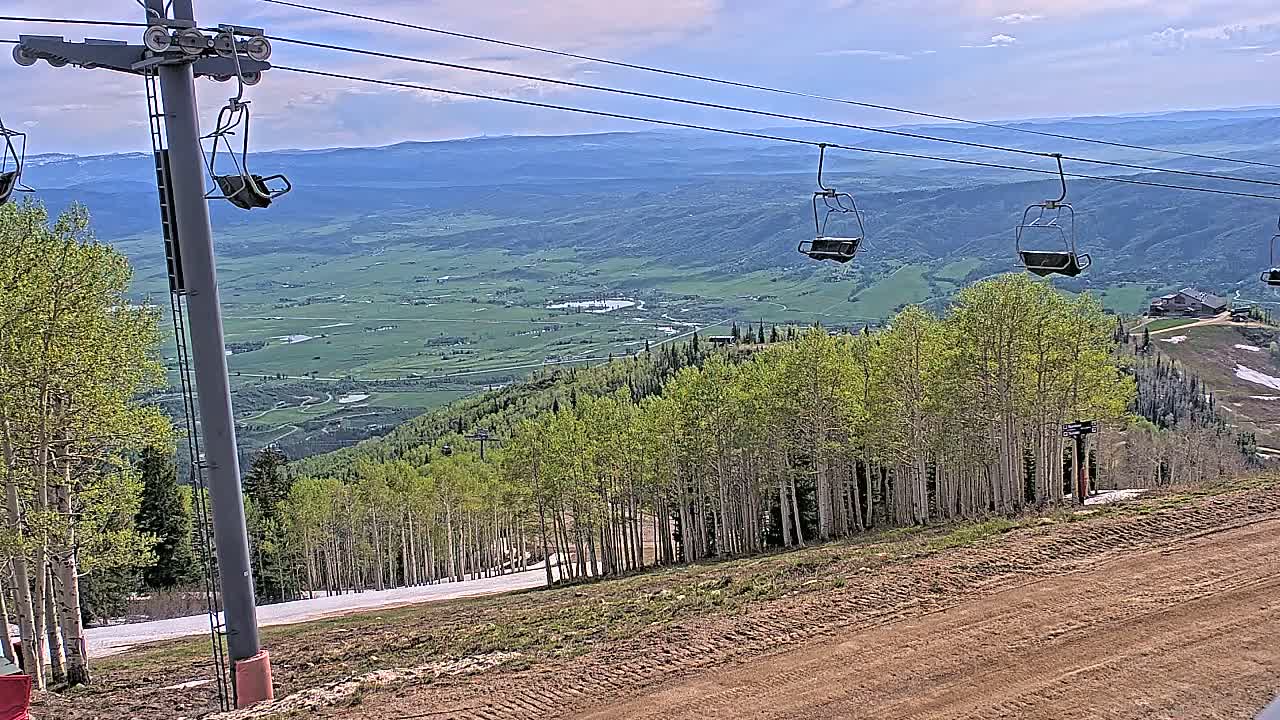 A chairlift ascends a mountain slope overlooking a vast green valley under a partly cloudy sky.
