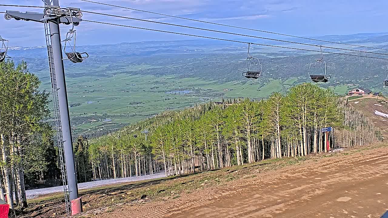 A ski lift with empty chairs overlooks a vast green valley under a mostly clear blue sky.