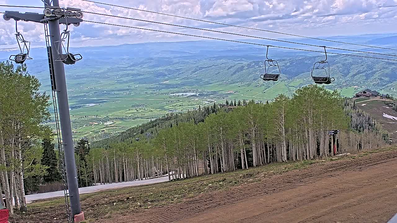 A mountaintop ski lift with two empty chairs overlooks a valley of green fields and forests under partly cloudy skies.
