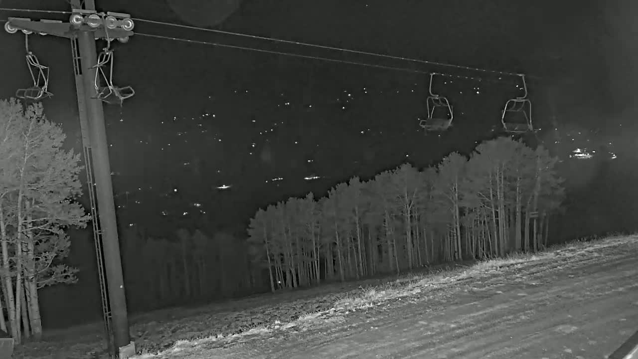 At night, empty ski lift chairs hang from a cable above a snow-covered landscape with a distant town illuminated by lights and a stand of trees in the foreground.
