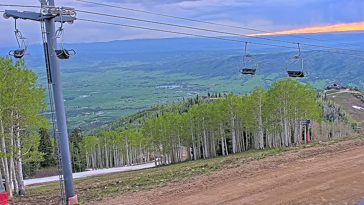 A ski lift with empty chairs overlooks a valley of green fields and aspen trees under a partly cloudy sunset sky.