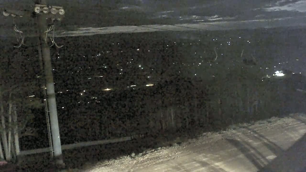 A nighttime view from a high vantage point shows a sparsely lit town below, with a snow-covered area in the distance under a cloudy sky.