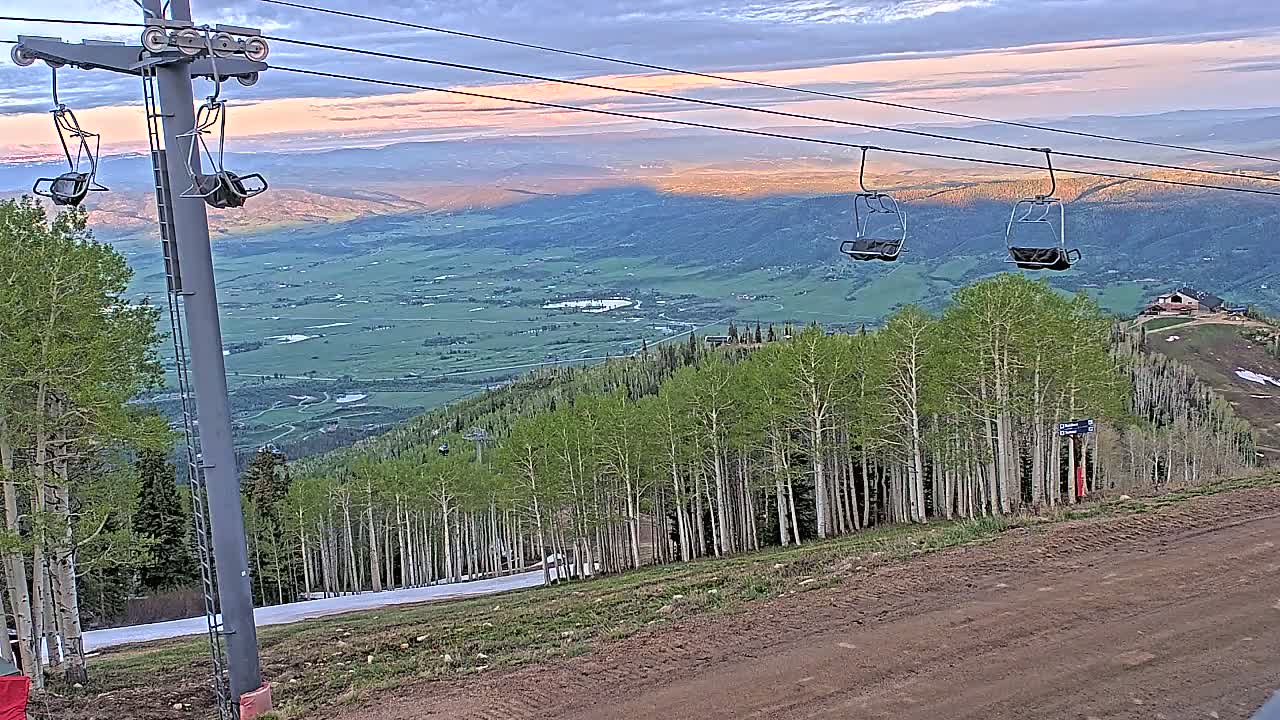 An empty chairlift overlooks a valley of green fields and aspen trees under a partly cloudy sunset sky.