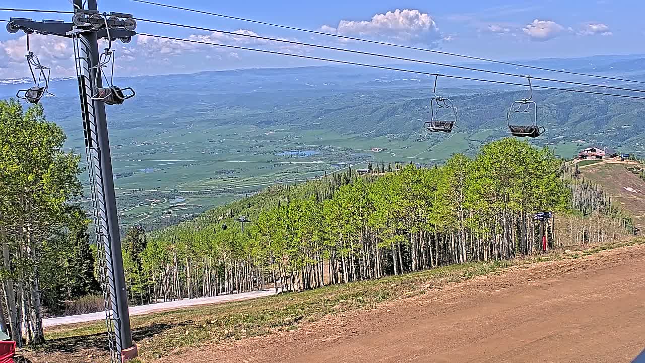 A ski lift with empty chairs overlooks a valley of green trees and fields under a partly cloudy sky.