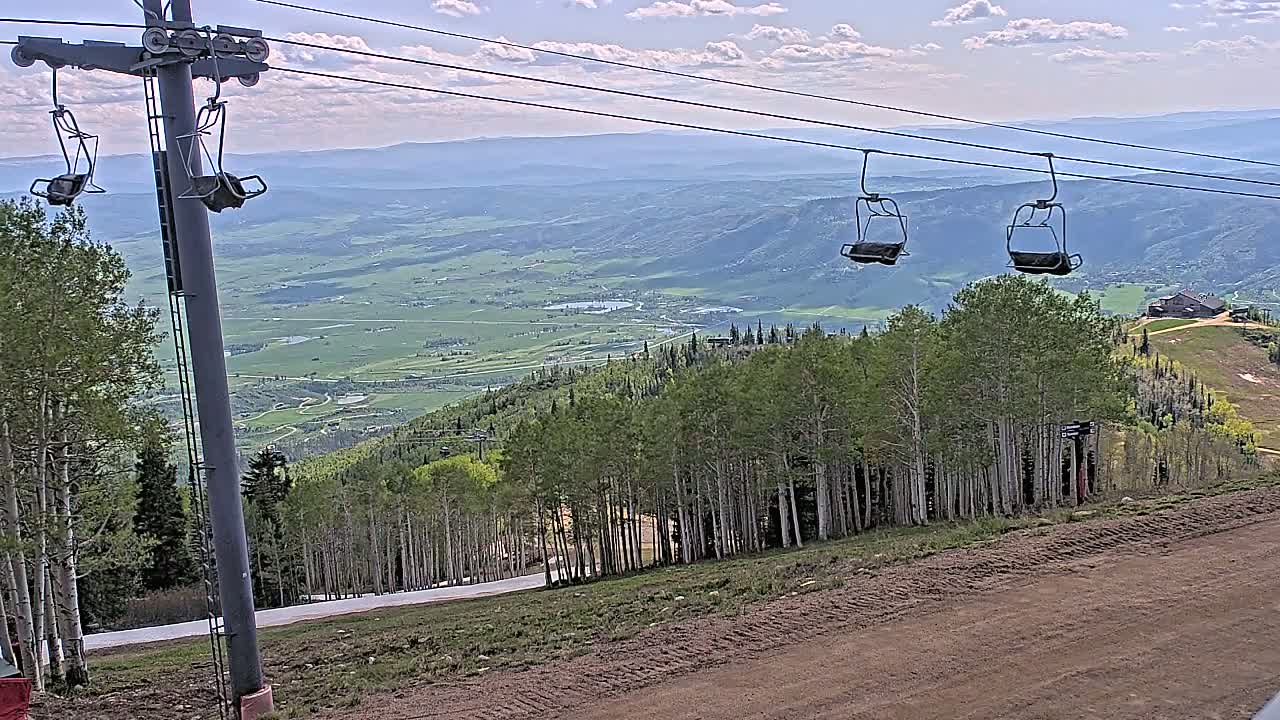 A ski lift with empty chairs overlooks a valley of green fields and a forest of aspen trees under partly cloudy skies.
