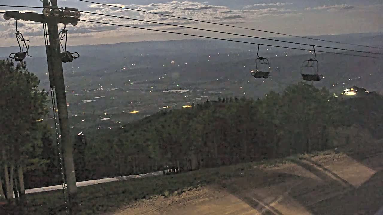 An empty chairlift hangs over a valley illuminated by distant lights under a partly cloudy night sky.