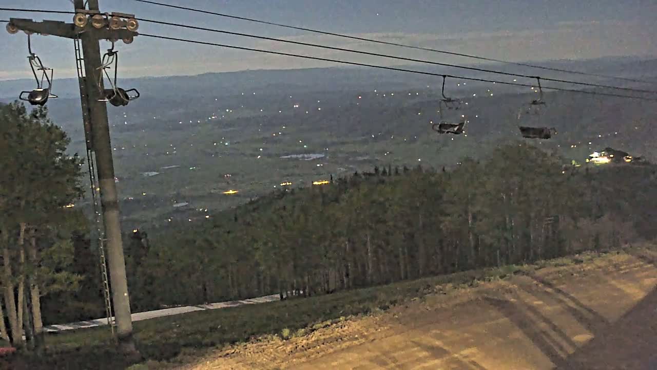 An empty chairlift at night overlooks a valley with distant lights under a dusky sky.