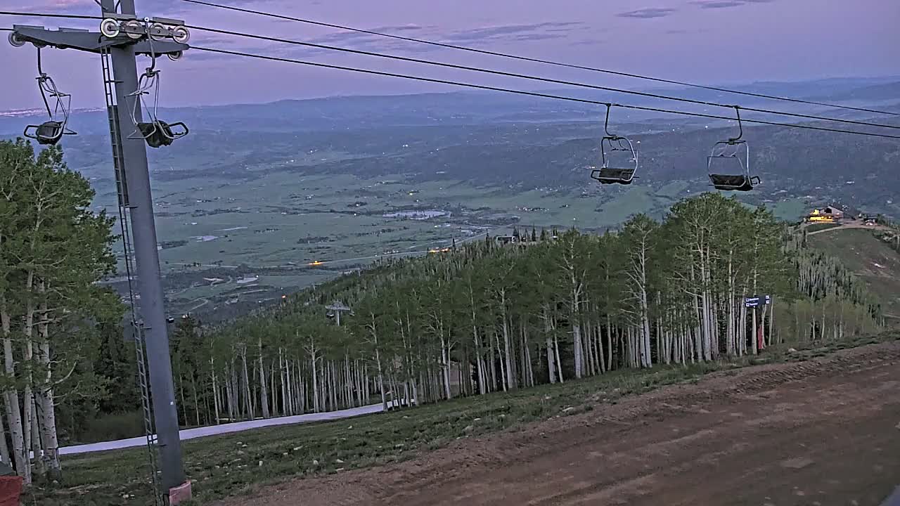 A chairlift ascends a mountain at dusk overlooking a vast valley, with a forest of slender trees below.