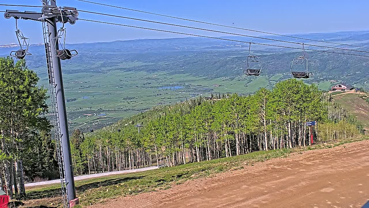 A ski lift with empty chairs overlooks a vast green valley under a clear blue sky.