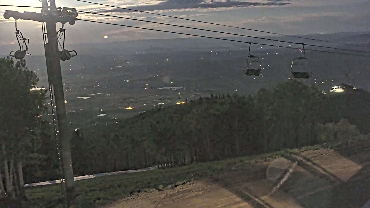 A chairlift with empty chairs overlooks a valley with scattered lights at twilight under a cloudy sky.