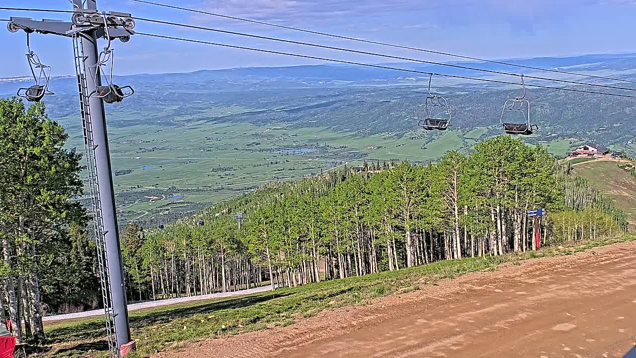A ski lift chair hangs above a dirt road leading to a lush green valley under a clear blue sky.