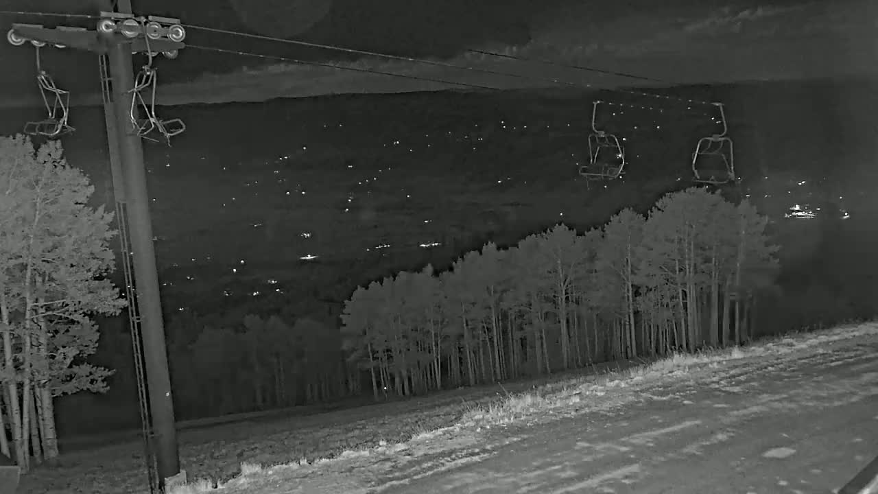 A nighttime view of a ski lift with empty chairs overlooking a town with scattered lights and a snow-covered slope below.