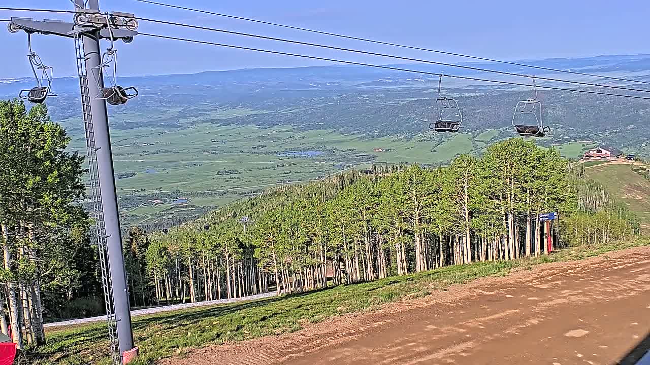 A ski lift with empty chairs overlooks a vast green valley under a clear blue sky.