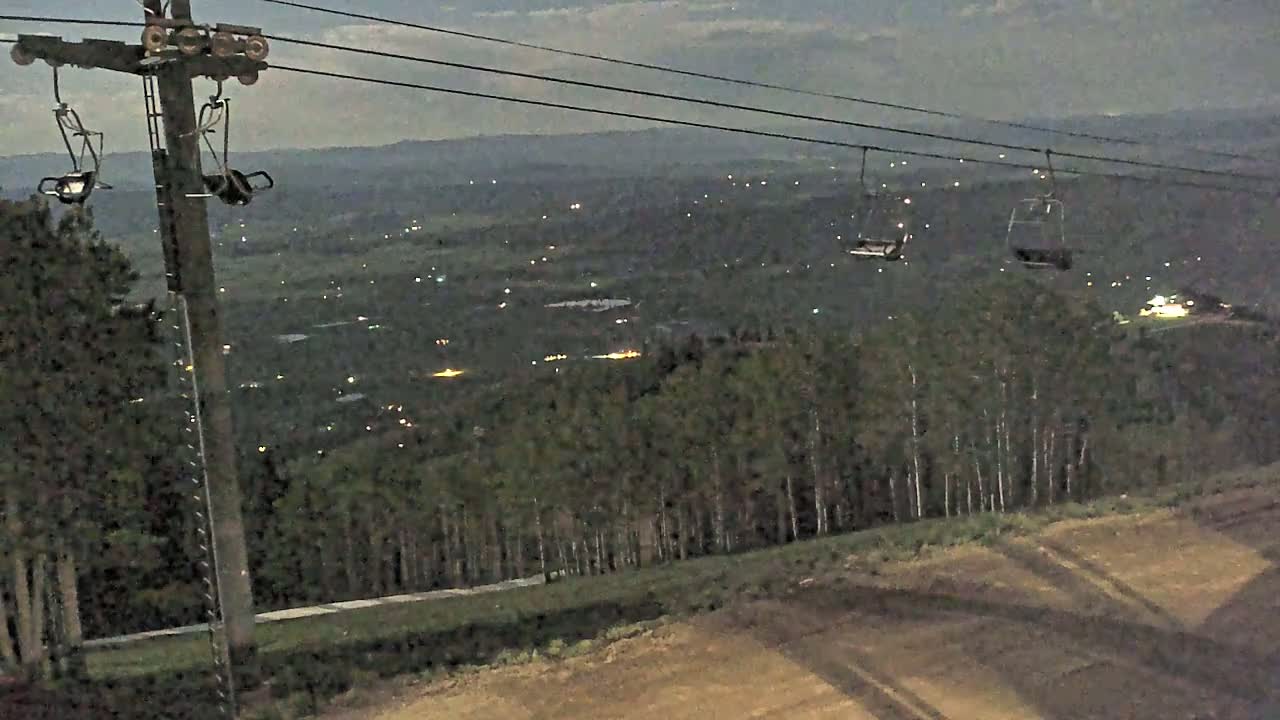 An empty chairlift ascends a hill overlooking a valley speckled with distant lights at dusk.