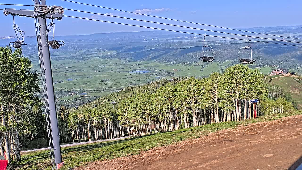 A chairlift ascends a mountainside of aspens on a sunny day, overlooking a vast green valley.