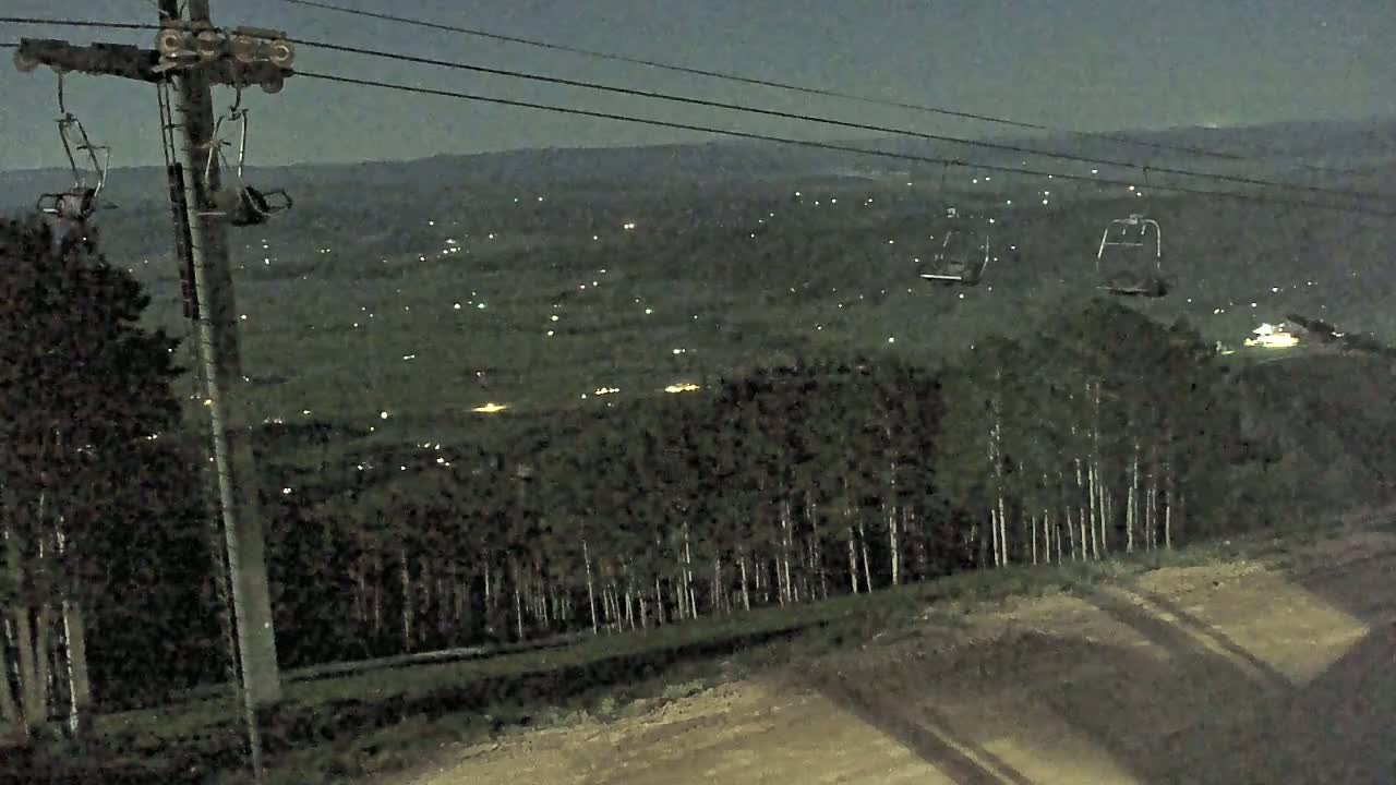 A ski lift at night overlooks a valley dotted with distant lights under a dark sky.