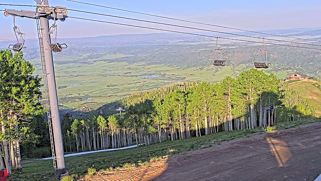 A chairlift ascends a mountain slope overlooking a vast valley under a clear, sunny sky.