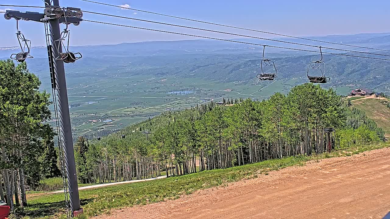 A ski lift chair hangs from cables overlooking a vast green valley under a clear blue sky.