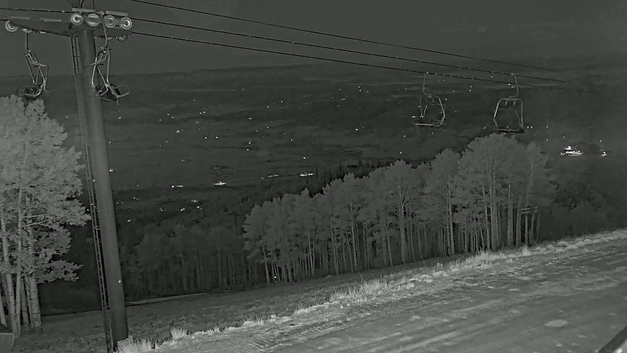 At night, a ski lift with empty chairs overlooks a snow-covered landscape dotted with trees and distant lights.